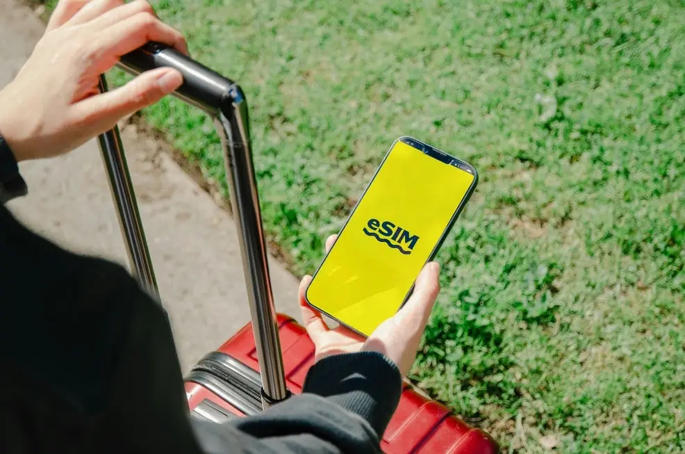 Person holding a smartphone with eSIM activation on the screen, ready for travel with a suitcase outdoors